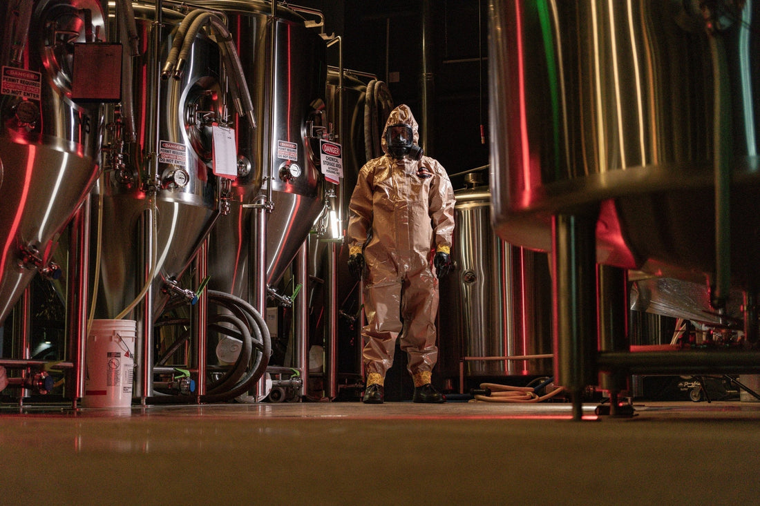 MIRA Safety hazmat suit and gas mask worn in an industrial setting with stainless steel tanks and warning signs. High-performance CBRN protective gear for hazardous environments and tactical preparedness.